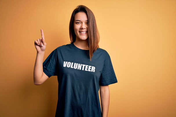 Молода красива брюнетка дівчина робить волонтерство в футболці з волонтерським словом повідомлення з великою посмішкою на обличчі, вказуючи пальцем на бік камери
. - Фото, зображення