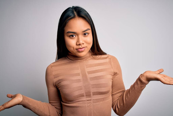 Young beautiful asian girl wearing casual turtleneck sweater standing over white background clueless and confused expression with arms and hands raised. Doubt concept. - Fotó, kép