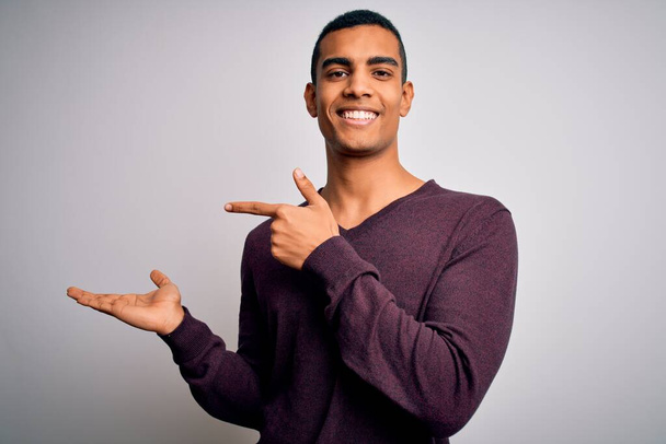 Junger gutaussehender afrikanisch-amerikanischer Mann in lässigem Pullover vor weißem Hintergrund erstaunt und lächelnd in die Kamera, während er mit der Hand präsentiert und mit dem Finger zeigt. - Foto, Bild