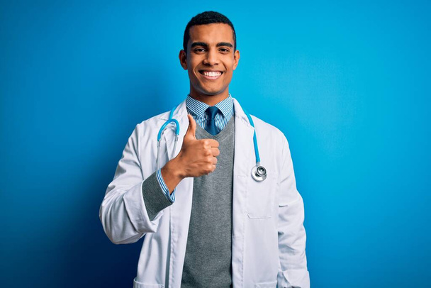 Przystojny, afrykański lekarz w płaszczu i stetoskopie na niebieskim tle, robiący gesty dłońmi w górę. Zatwierdzanie ekspresji patrząc w aparat pokazujący sukces. - Zdjęcie, obraz
