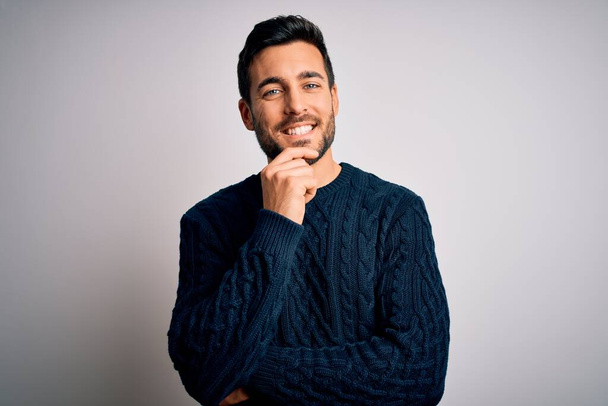 Young handsome man with beard wearing casual sweater standing over white background looking confident at the camera with smile with crossed arms and hand raised on chin. Thinking positive. - Photo, Image