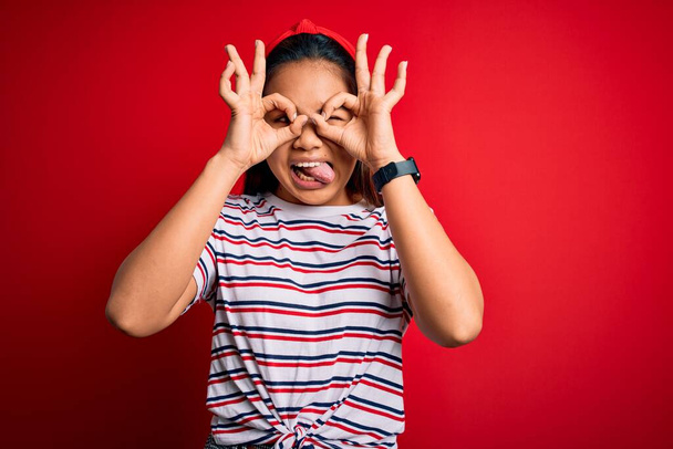 Young beautiful asian girl wearing casual striped t-shirt over isolated red background doing ok gesture like binoculars sticking tongue out, eyes looking through fingers. Crazy expression. - Foto, Bild