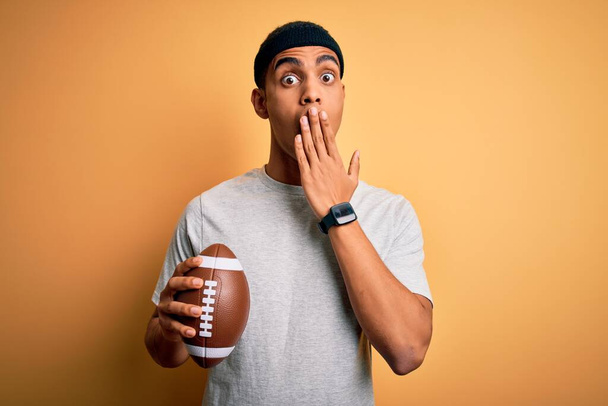Young handsome african american man holding rugby ball over isolated yellow background cover mouth with hand shocked with shame for mistake, expression of fear, scared in silence, secret concept - Foto, Imagem