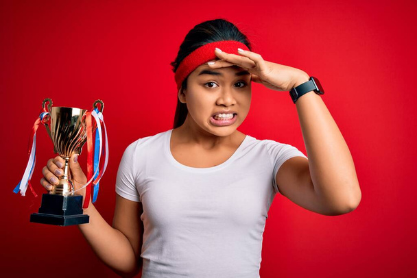 Jonge kampioen aziatische meisje winnende trofee beker staande over geïsoleerde rode achtergrond gestrest met de hand op het hoofd, geschokt door schaamte en verrassing gezicht, boos en gefrustreerd. Angst en overstuur voor fouten. - Foto, afbeelding