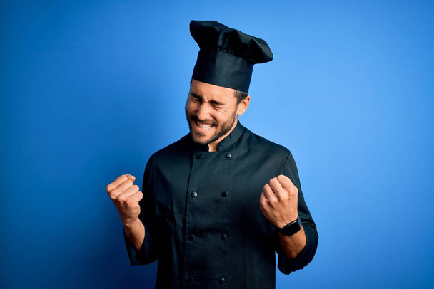 Jeune beau chef homme avec barbe portant uniforme de cuisinière et chapeau sur fond bleu excité pour le succès avec les bras levés et les yeux fermés célébrant la victoire en souriant. Concept gagnant
. - Photo, image