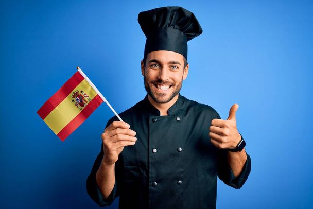 Νεαρός όμορφος μάγειρας με γενειάδα φορώντας στολή κρατώντας ισπανική σημαία ευτυχισμένη με μεγάλο χαμόγελο κάνει εντάξει σημάδι, αντίχειρας επάνω με τα δάχτυλα, εξαιρετική πινακίδα - Φωτογραφία, εικόνα