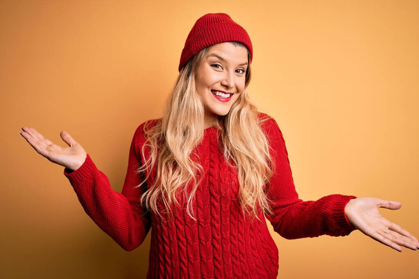 Mladá krásná blondýna žena na sobě ležérní svetr a vlněná čepice přes bílé pozadí s úsměvem ukazující obě ruce otevřené dlaně, prezentační a reklamní srovnání a rovnováhu - Fotografie, Obrázek