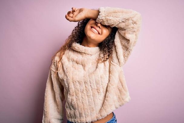 Jonge mooie vrouw met krullend haar dragen casual trui staan over roze achtergrond bedekken ogen met arm glimlachend vrolijk en grappig. Blind concept. - Foto, afbeelding