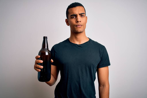 Joven hombre afroamericano guapo bebiendo botella de cerveza sobre fondo blanco con una expresión segura en la cara inteligente pensando en serio
 - Foto, imagen