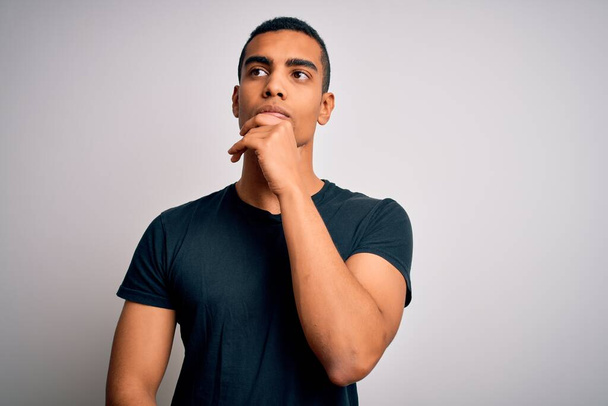 Young handsome african american man wearing casual t-shirt standing over white background with hand on chin thinking about question, pensive expression. Smiling and thoughtful face. Doubt concept. - Zdjęcie, obraz