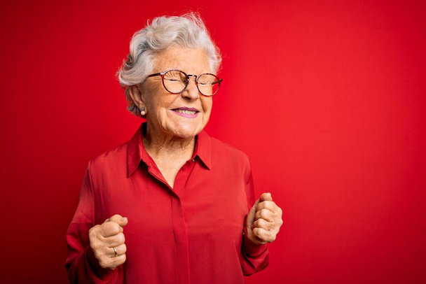 Starsza piękna siwowłosa kobieta w luźnej koszuli i okularach na czerwonym tle podekscytowana sukcesem z podniesionymi ramionami i zamkniętymi oczami świętującymi zwycięstwo uśmiechnięta. Koncepcja zwycięzcy. - Zdjęcie, obraz