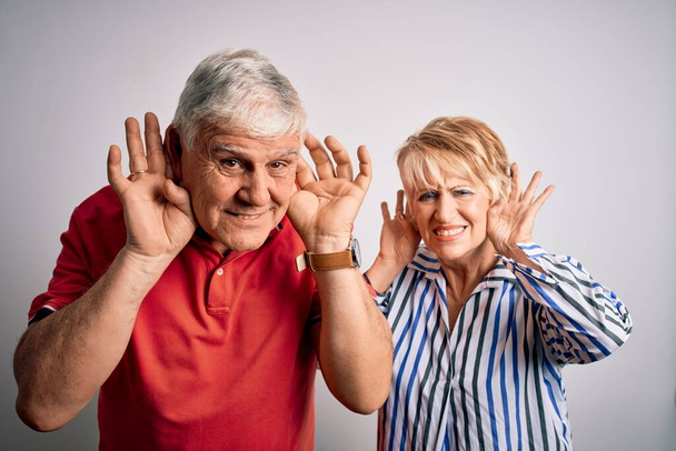 Senior beau couple debout ensemble sur fond blanc isolé Essayant d'entendre les deux mains sur le geste de l'oreille, curieux de commérages. Problème auditif, sourd
 - Photo, image