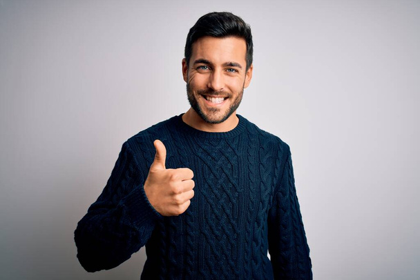 Young handsome man with beard wearing casual sweater standing over white background doing happy thumbs up gesture with hand. Approving expression looking at the camera showing success. - Photo, image