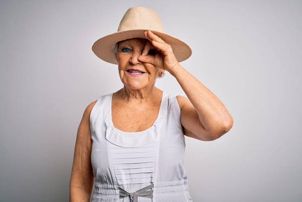 Senior krásná šedovlasá žena na dovolené na sobě ležérní letní šaty a klobouk dělá v pořádku gesto s ručním úsměvem, oko dívá přes prsty se šťastnou tváří. - Fotografie, Obrázek