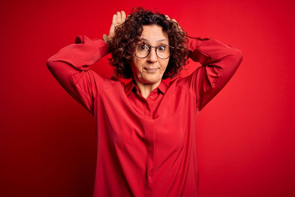 Middle age beautiful curly hair woman wearing casual shirt and glasses over red background Doing bunny ears gesture with hands palms looking cynical and skeptical. Easter rabbit concept. - Photo, Image
