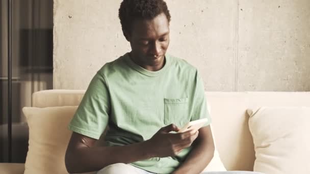 A calm african american man in casual clothes is using his smartphone while sitting on the sofa at home - Πλάνα, βίντεο