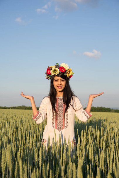schöne lächelnde indische Mädchen in traditioneller ukrainischer Kleidung und handgefertigten Blumenkranz stehen in Roggen Feld mit den Händen in namaste Grußgeste, ethnische junge Frau auf Himmel Natur Hintergrund - Foto, Bild