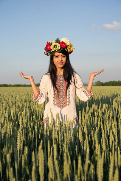 Beautiful smiling indian girl in traditional Ukrainian clothes and handmade flowery wreath stand in rye field with hands in namaste greeting gesture, ethnic young woman on sky nature background - Photo, Image