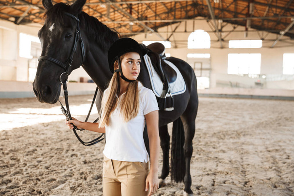 Image of concentrated young woman wearing hat with horse in countryside in corral on training area. - Fotoğraf, Görsel