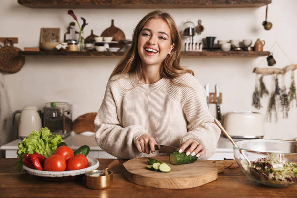 Image of pleased cute woman wearing casual clothes laughing and making lunch in cozy kitchen - Foto, imagen
