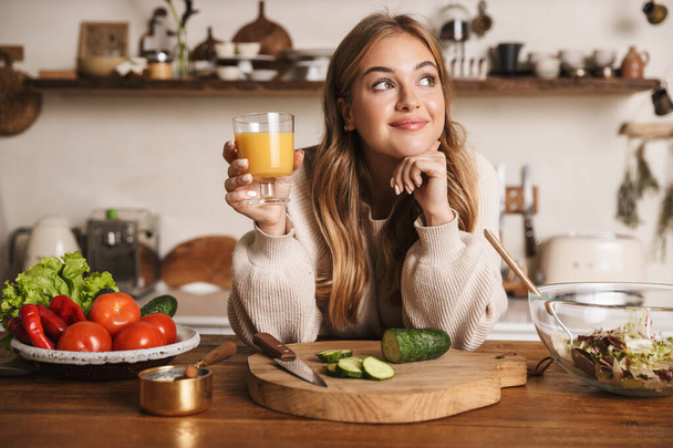 Image of pleased cute woman wearing casual clothes drinking juice while making lunch in cozy kitchen - Фото, зображення
