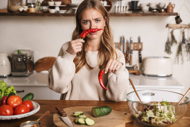 Image of sad cute woman wearing casual clothes grimacing while cooking dinner in cozy kitchen - Foto, Bild