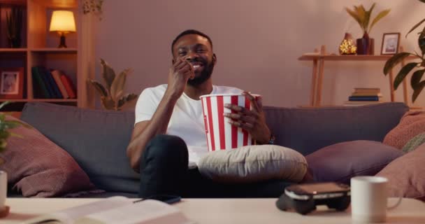 Front view of relaxed handsome guy watching funny comedy film and laughing. Smiling young man eating popcorn from paper bowl while sitting on couch in front of TV.Indoors. - Кадры, видео