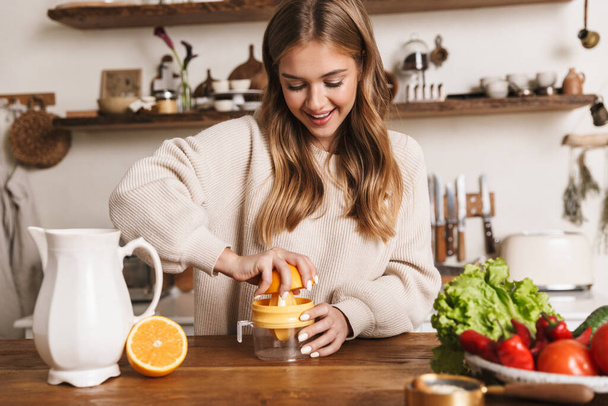 Image of happy caucasian woman wearing casual clothes making orange juice and smiling in cozy kitchen - Fotoğraf, Görsel