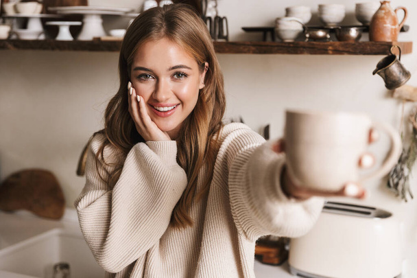 Image of beautiful funny woman wearing casual clothes drinking coffee and looking at camera in cozy kitchen - Foto, Imagen