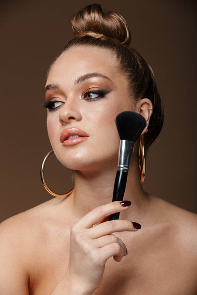 Close up beauty portrait of an attractive blonde young topless woman isolated over background, wearing accessories, holding makeup brush at her face - Φωτογραφία, εικόνα