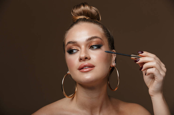Close up beauty portrait of an attractive blonde young topless woman isolated over background, wearing accessories, doing makeup with mascara - Φωτογραφία, εικόνα