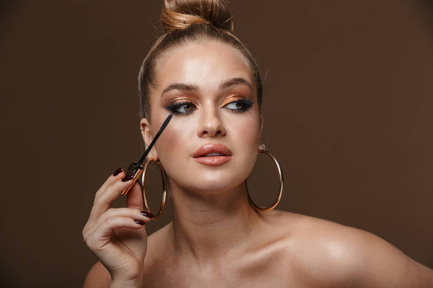 Close up beauty portrait of an attractive blonde young topless woman isolated over background, wearing accessories, doing makeup with mascara - Zdjęcie, obraz