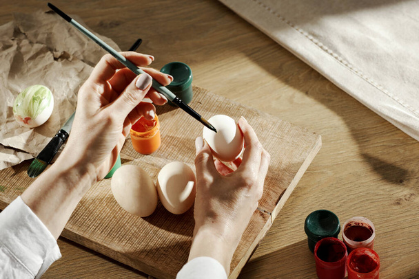 Kobiecy ręka maluję wielkanocny jajko na drewniany stół na wiosenny dzień. Czas dekoracji wielkanocnej.Kolorowe jaja i farby na drewnianej desce. - Zdjęcie, obraz