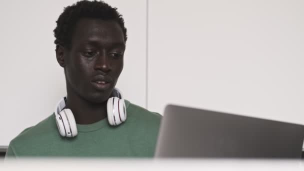 Uma vista recortada de um jovem americano africano concentrado e calmo está usando seu computador portátil prateado sentado à mesa na sala de estar
 - Filmagem, Vídeo