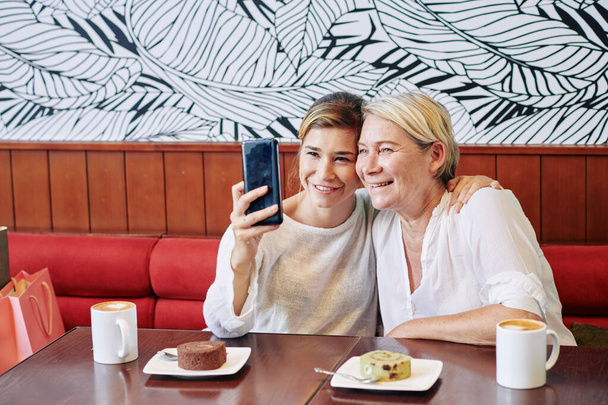 Hugging young pretty woman having coffee with her mother and photographing on smatphone - 写真・画像