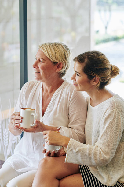 Mother and daughter drinking morning cappuccino and looking outdoors through big window - Foto, Imagen