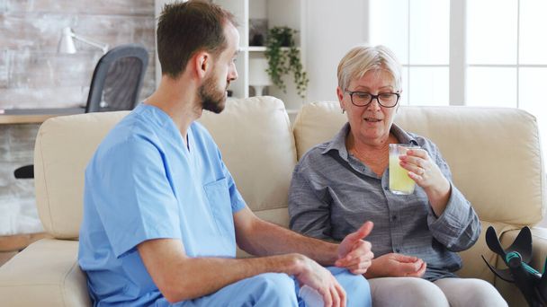 Enfermeira do sexo masculino sentado no sofá com a mulher sênior dando seu tratamento médico em casa de repouso
 - Foto, Imagem