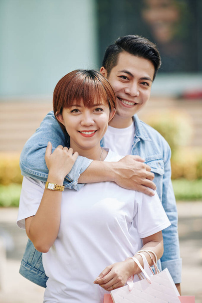 Happy young Asian man hugging his pretty girlfriend with many shopping bags from behind when that standing outdoors - Photo, Image