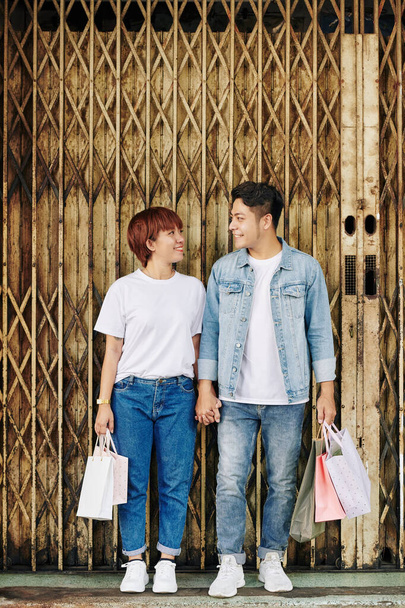 Веселая молодая пара влюблённая, держащая сумки с покупками и смотрящая друг на друга
 - Фото, изображение