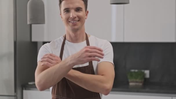 Glimlachende jongeman met een bruin schort kruist zijn handen terwijl hij in een zelfverzekerde houding in de keuken staat - Video