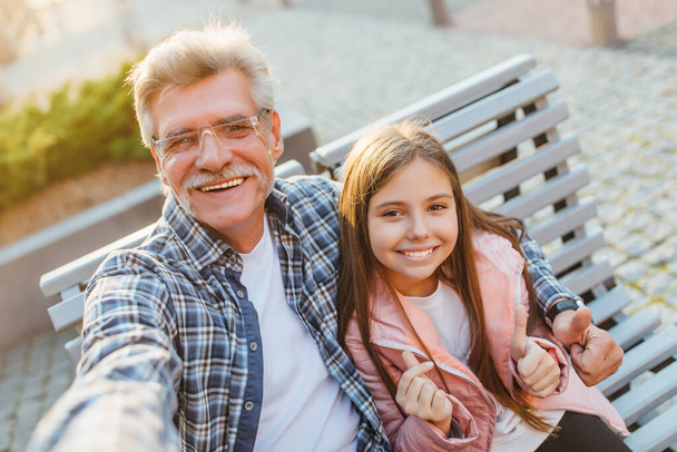 Photo of a grandfather and granddaughter sitting on a bench and make a selfie. - Foto, Imagen