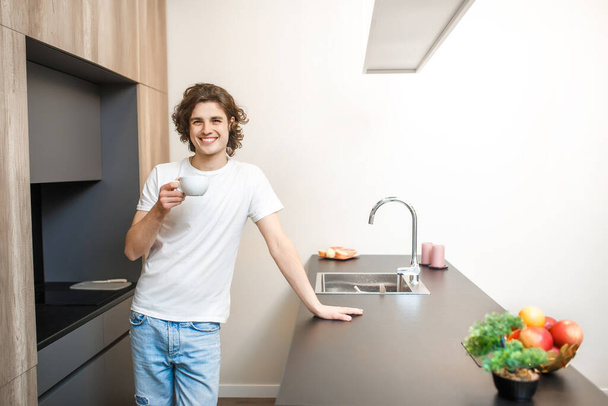 Portrait of handsome man in casual white t-shirt drinking morning coffee in kitchen, healthy time. - Foto, Bild