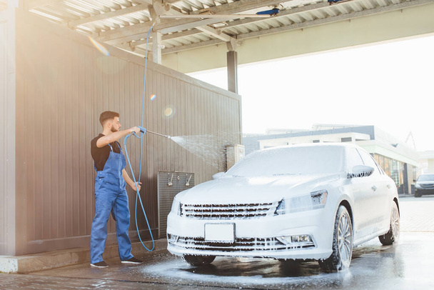 Professional washer in blue uniform washing luxury car with water gun on an open air car wash. - Foto, afbeelding