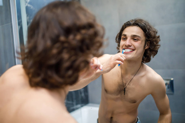 Health care, dental hygiene, people and beauty concept, smiling young man with toothbrush cleaning teeth and looking to the mirror at bathroom. - Φωτογραφία, εικόνα