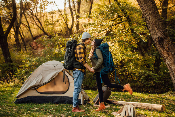 Young beautiful couple with hiking backpack kissing in the forest near tent. Attractive woman and handsome man relaxing together in nature. - Фото, зображення
