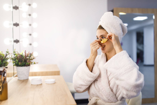 Beautiful girl in a white bathrobe and with a towel on her head puts patches under her eyes in front of a mirror. - Foto, afbeelding