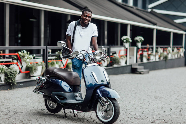 Handsome man riding a scooter on city roads. Summer potrait of male wearing a white shirt sitting near  retro motorbike. - Fotó, kép