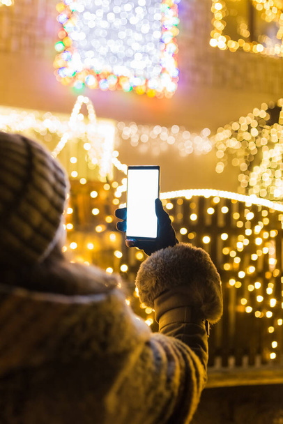 Rear view of young female holding blank screen mobile phone against house decorated with christmas lights. - Foto, Imagem