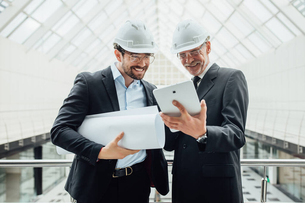 Dwóch młodych biznesmenów lub pracownik budowlany w kasku stoi w białym budynku, trzymając kartkę papieru, rzucając. - Zdjęcie, obraz