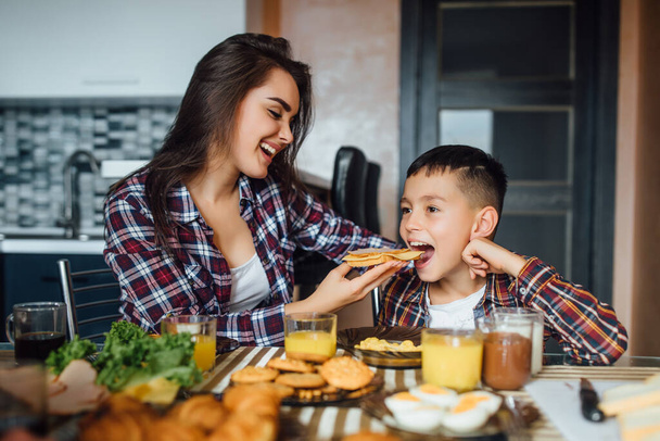 Cheerful brunette mother feeding her son sandwich with cheese during breakfast at home. - Valokuva, kuva